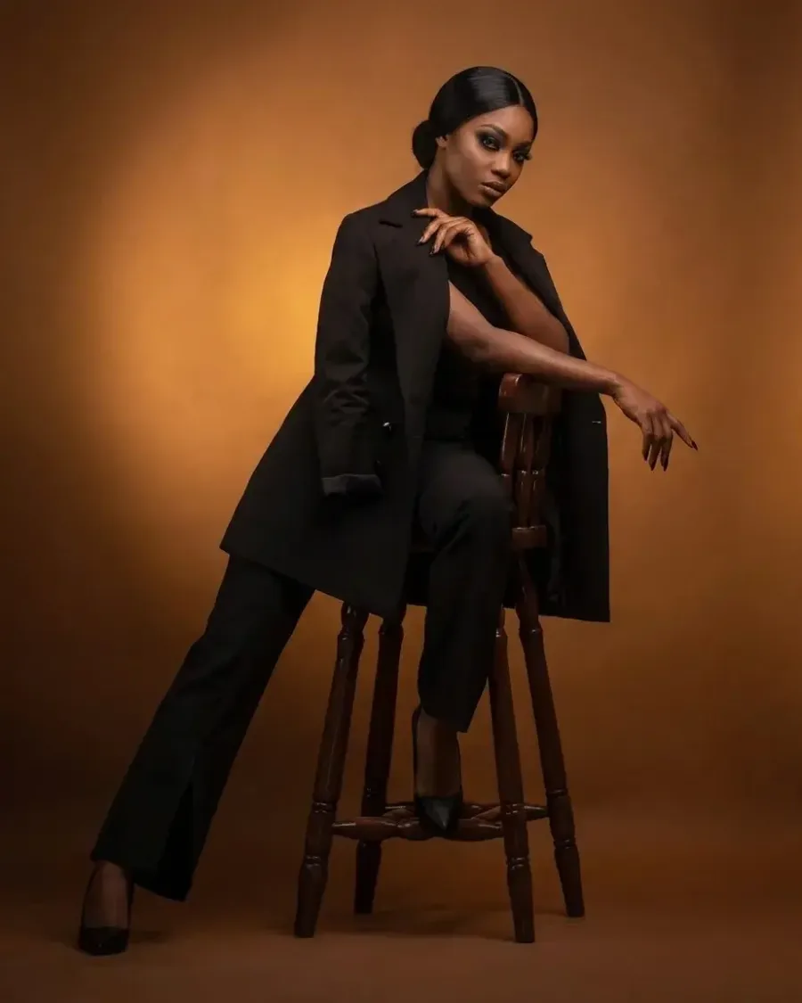 A woman in a long black coat and trousers seated on a wooden stool against a warm amber backdrop