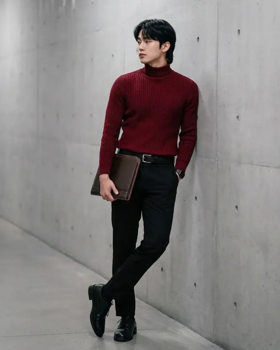 A man in a burgundy turtleneck and black trousers against a concrete wall