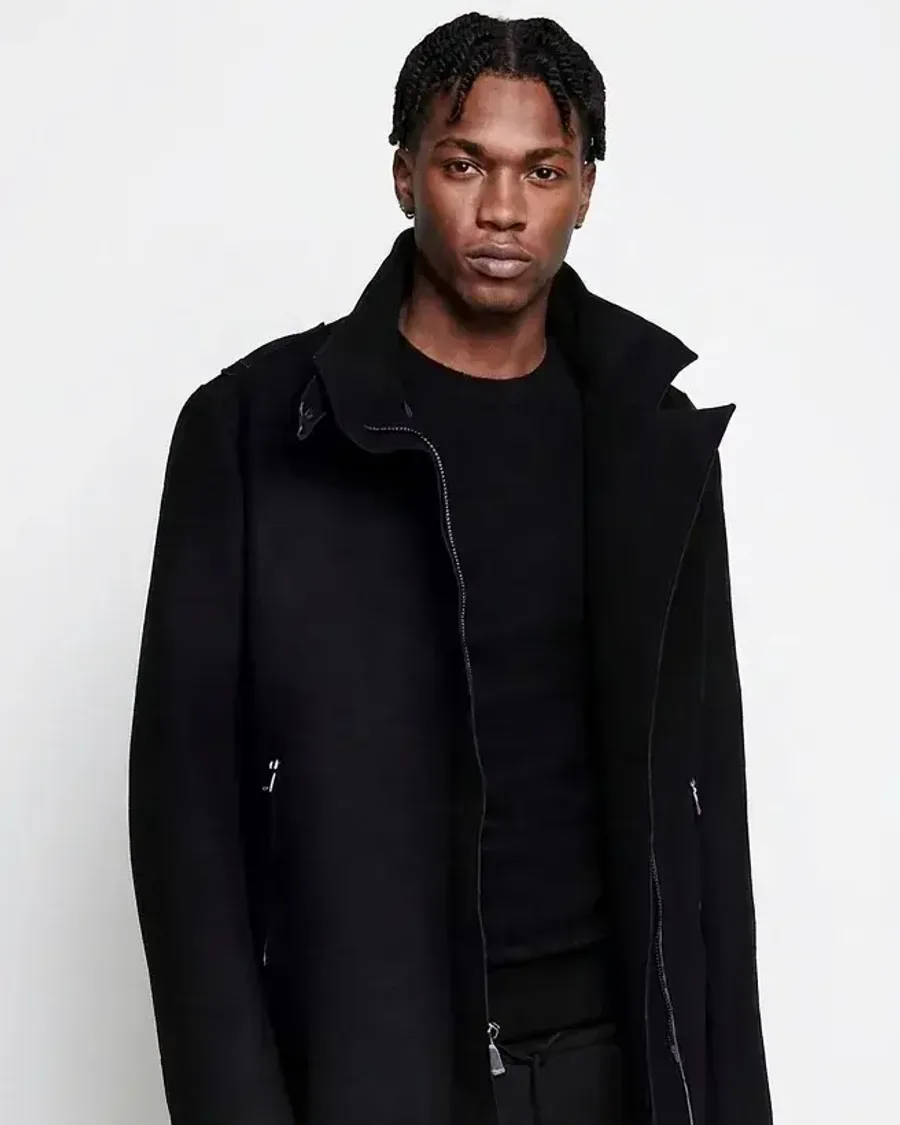 A man in a long black overcoat over a black knit against a white studio wall
