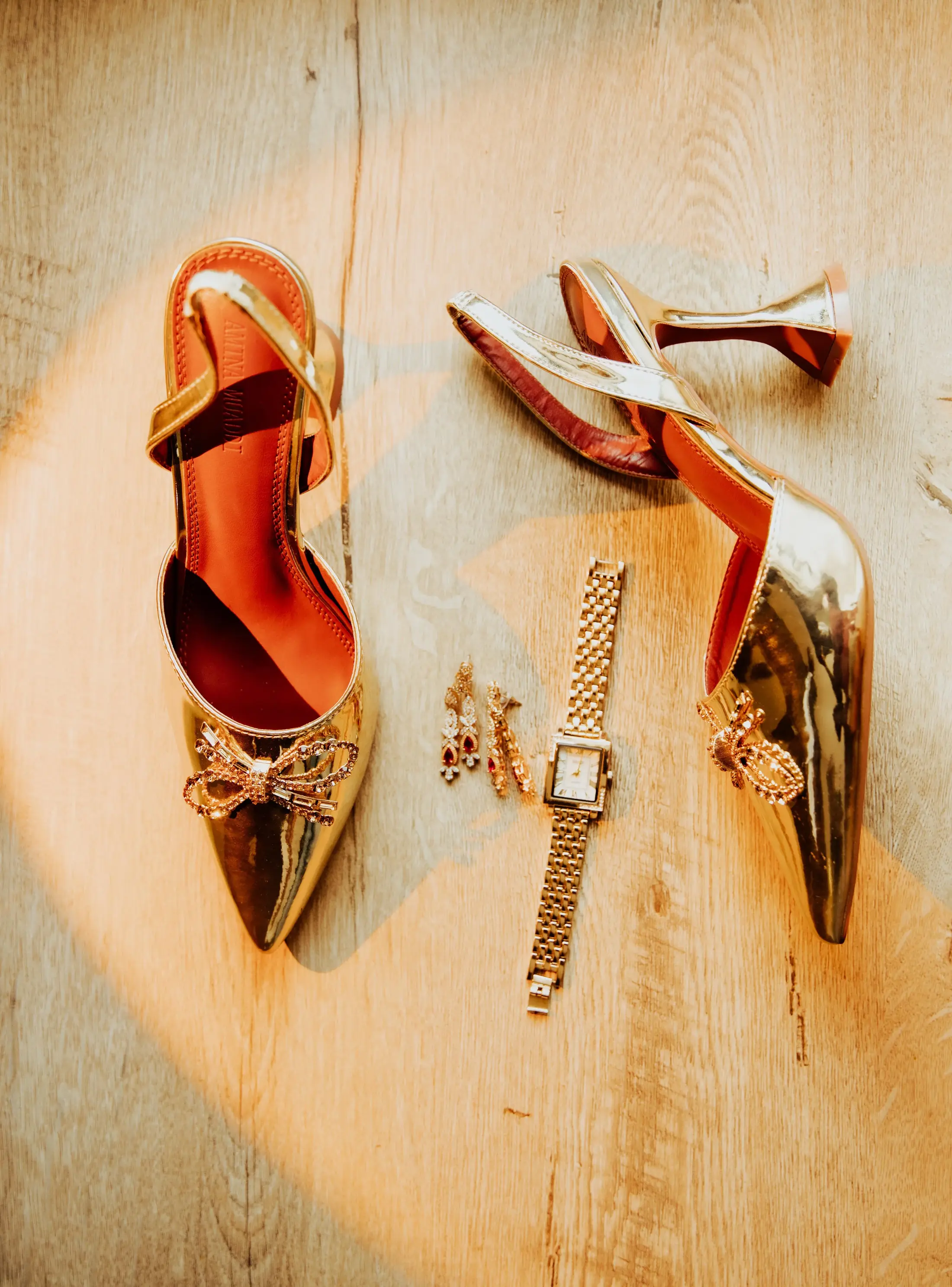 Gold heels, a wristwatch, and earrings on a sunlit wooden floor